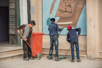 Three boys are sweeping the street. In the background is an orange dustbin and a picture hanging on the outside of a building.