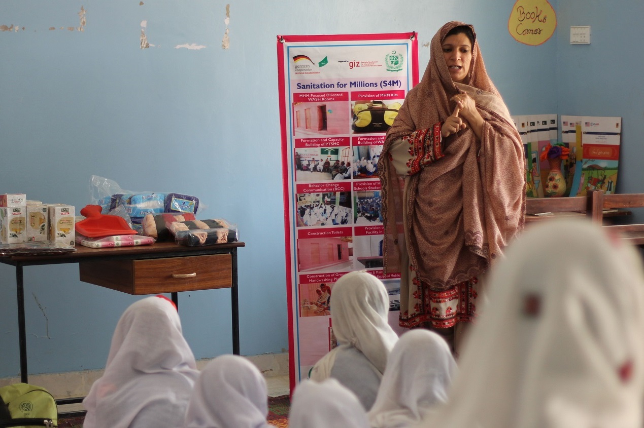 A women standing in front of a girls class and explaining something with a poster. Next to her are some Menstrual Hygiene Kits.