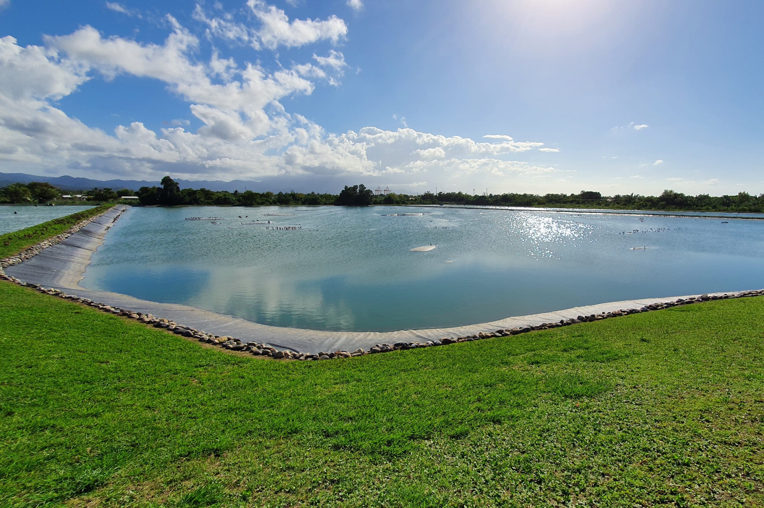 Ein blauer, künstlich angelegter Teich mit ein wenig Grünfläche im Vordergrund und dem blauen Himmel im Hintergrund.