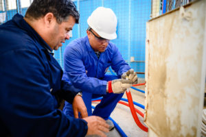Zwei Männer in blauen Overalls knien vor einer Wand und reparieren einen Schlauch.