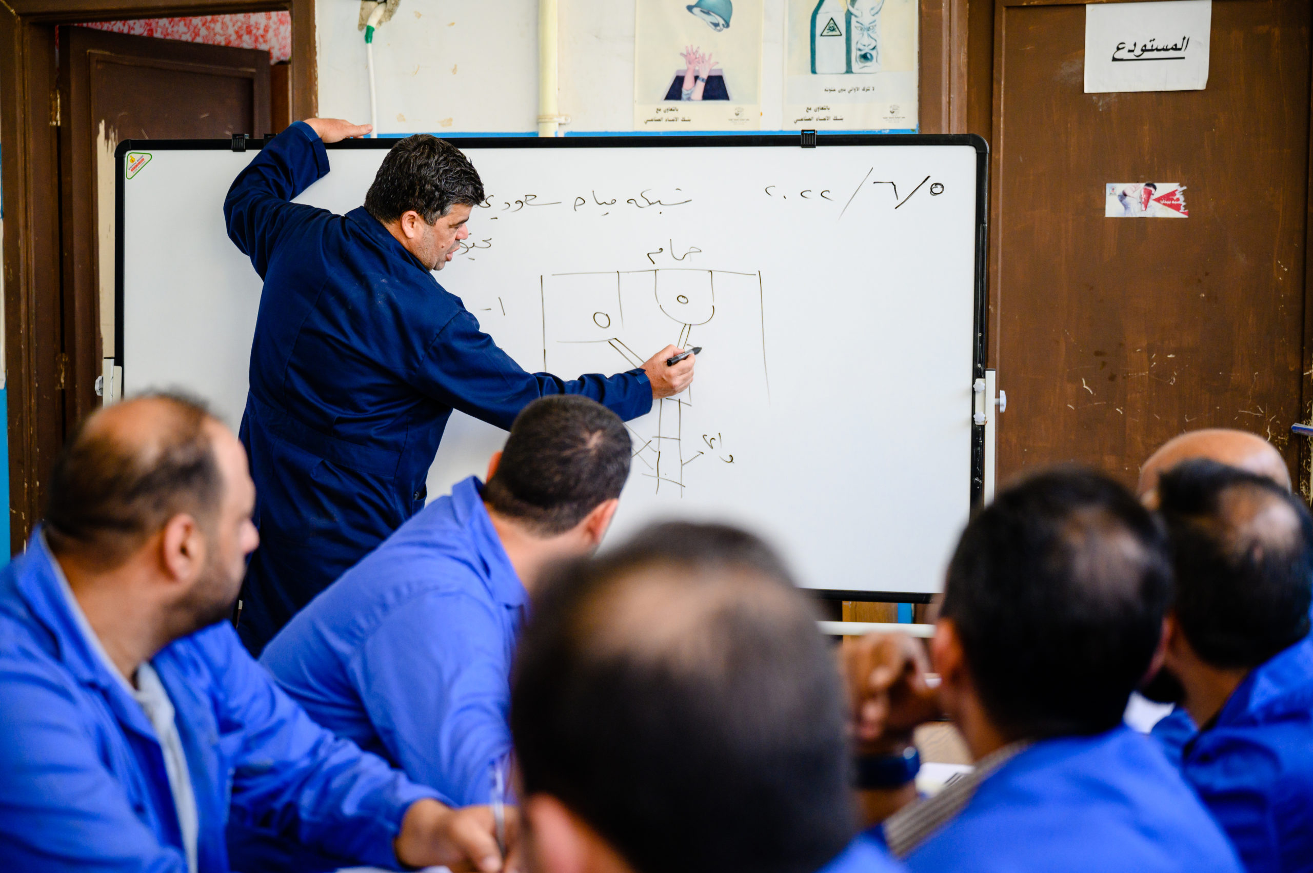 A man wearing a blue overall writes someting on a chalk board. Other men sitting in the classroom.