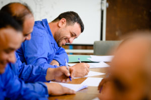 Drei Männer in blauen Overalls sitzen in einem Übungsraum und schreiben lächelnd etwas auf ein Blatt Papier.