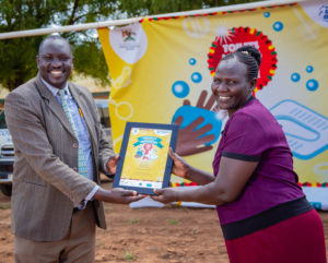 Ein Mann übergibt einer Frau eine Urkunde. Die beiden lachen in die Kamera und stehen draußen vor einem Plakat anlässlich des Wettbewerbs Toilets Making the Grade in Schulen in Uganda.