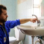 A tradesman in blue overalls kneels in front of a wash basin and checks whether the water tap is working.
