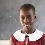 An Ugandan schoolgirl stands in front of a blackboard and smiles at the camera.