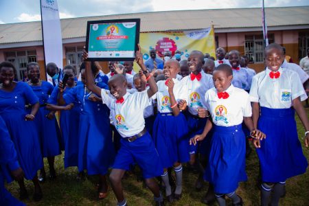 Ugandan school children wearing white-blue school uniforms are running towards the camera. One of the children holds up a certificate.