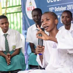 Ein Junge aus Uganda sitzt auf einer Bühne und spricht etwas in ein Mikrofon. Im Hintergrund sitzen weitere Schüler und Schülerinnen in ihren weiß-türkisen Schulunformen.