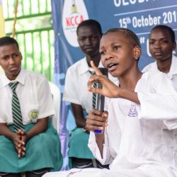 GIZ WASH-54 (1) Ein Junge aus Uganda sitzt auf einer Bühne und spricht etwas in ein Mikrofon. Im Hintergrund sitzen weitere Schüler und Schülerinnen in ihren weiß-türkisen Schulunformen.